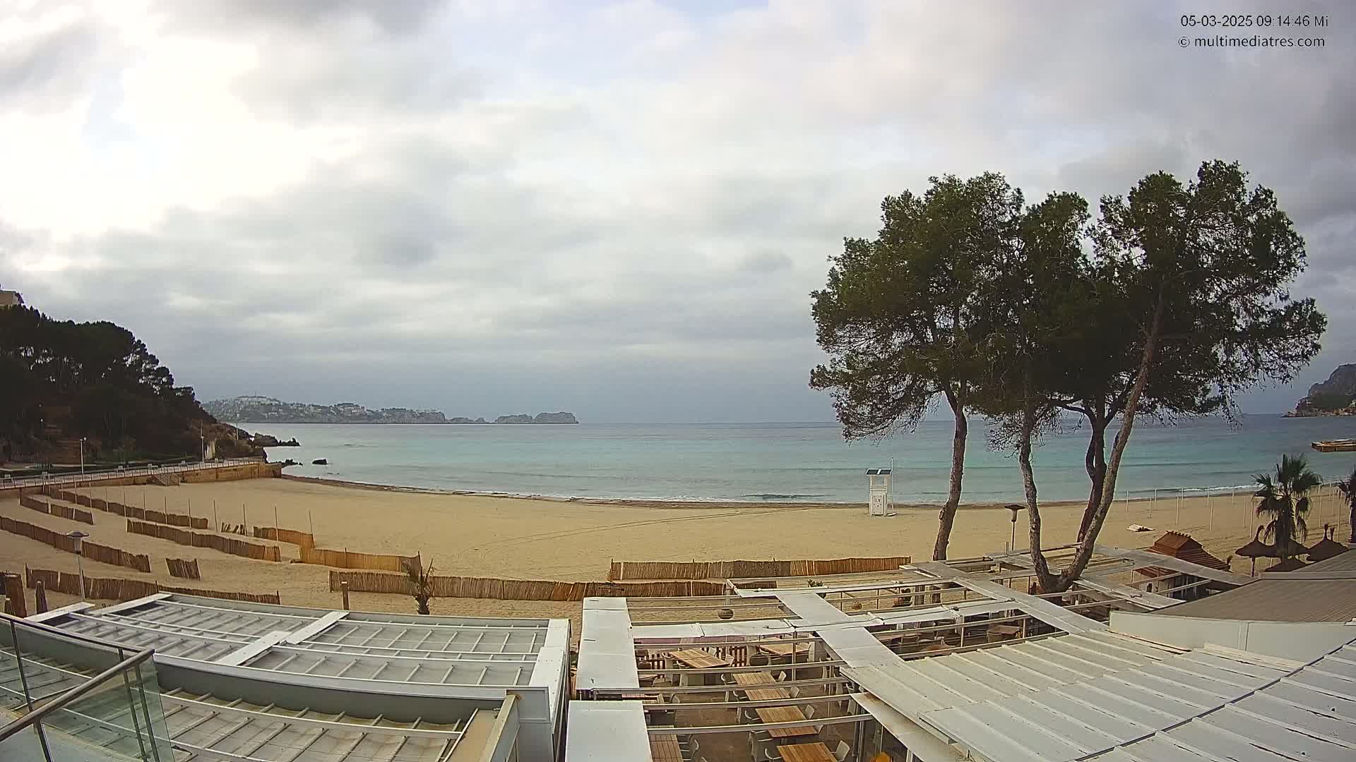 A sandy beach with calm, turquoise water stretches beneath an overcast sky, overlooked by several pine trees and a beachfront restaurant with retractable roofing.