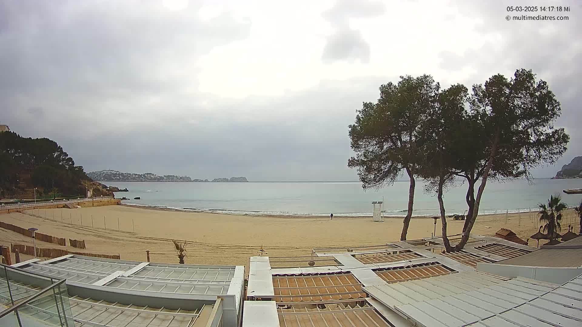 A mostly cloudy day shows a sandy beach with calm water, a lifeguard stand, and several buildings with covered rooftops overlooking the ocean.