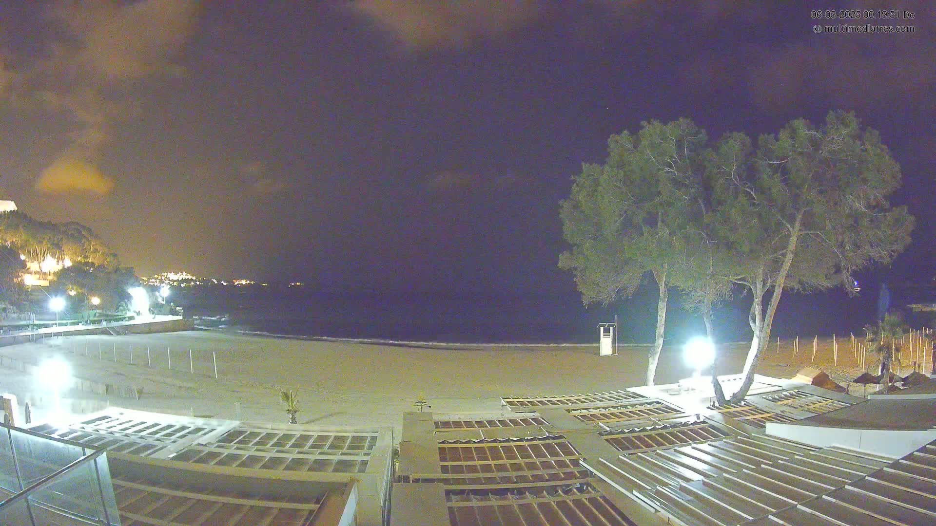 A nighttime view of a mostly empty sandy beach with several closed beach umbrellas and two tall, slender trees under a partly cloudy sky.