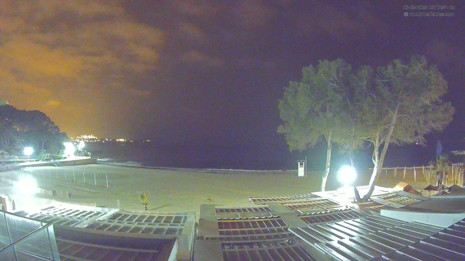 A nighttime view of a sandy beach with calm waves under a cloudy, amber sky, illuminated by lights near two trees and beach structures.