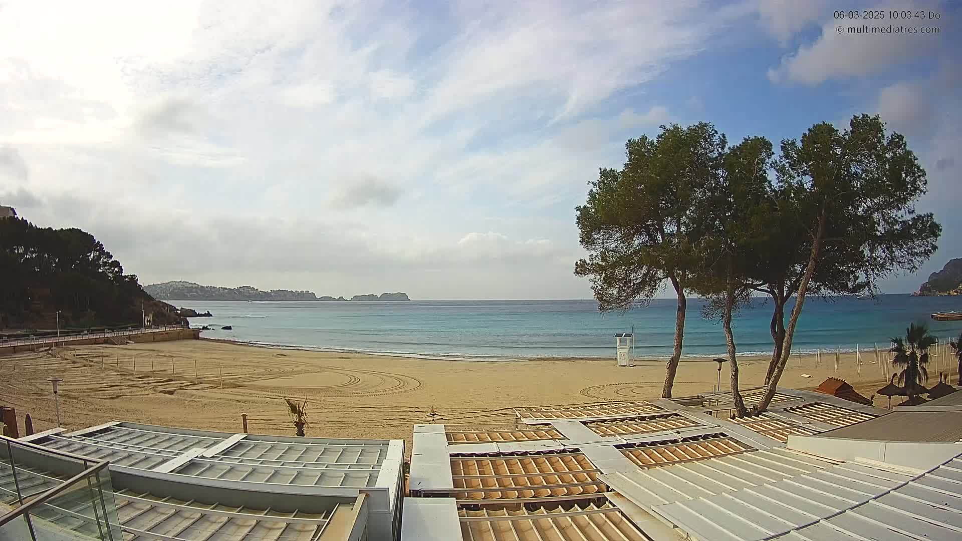 A mostly sunny day reveals a sandy beach with calm, turquoise water, a few trees, and several closed beachside structures.