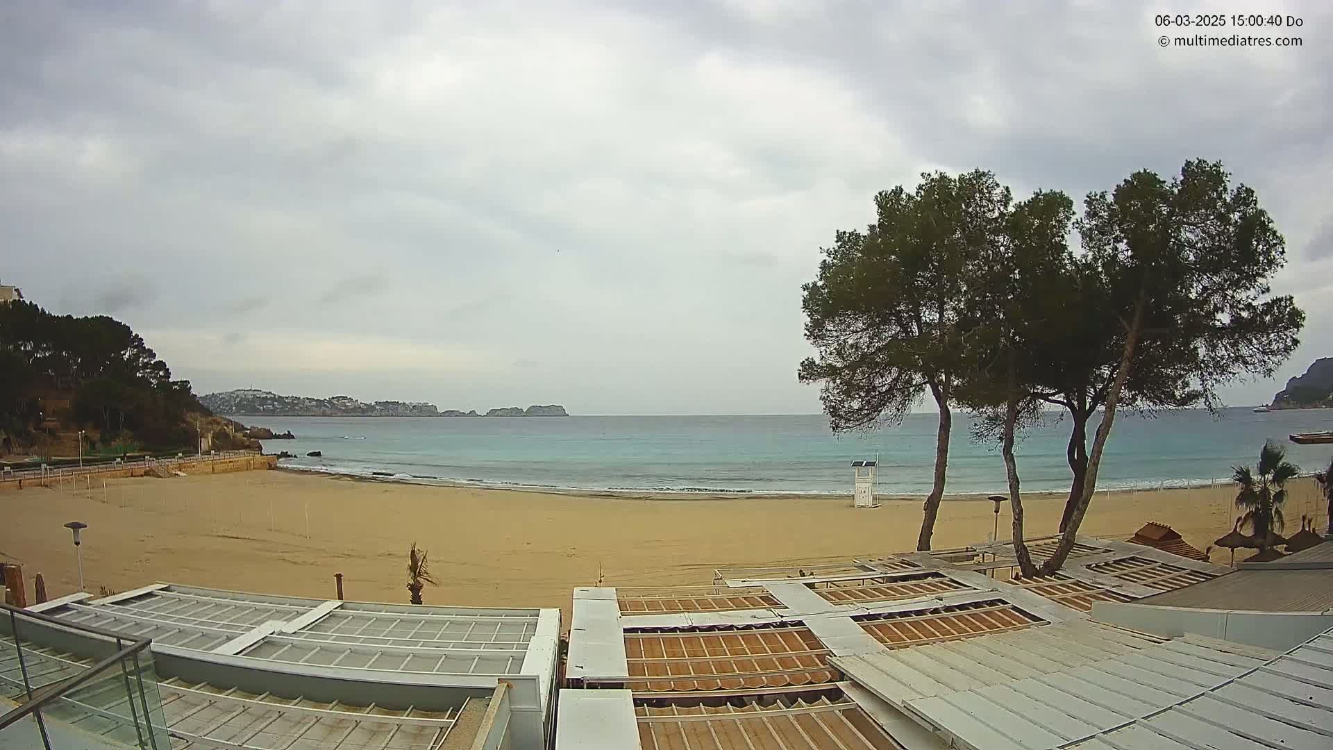 A sandy beach with calm, teal water stretches to a distant coastline under an overcast sky, with several pine trees and beach structures visible in the foreground.
