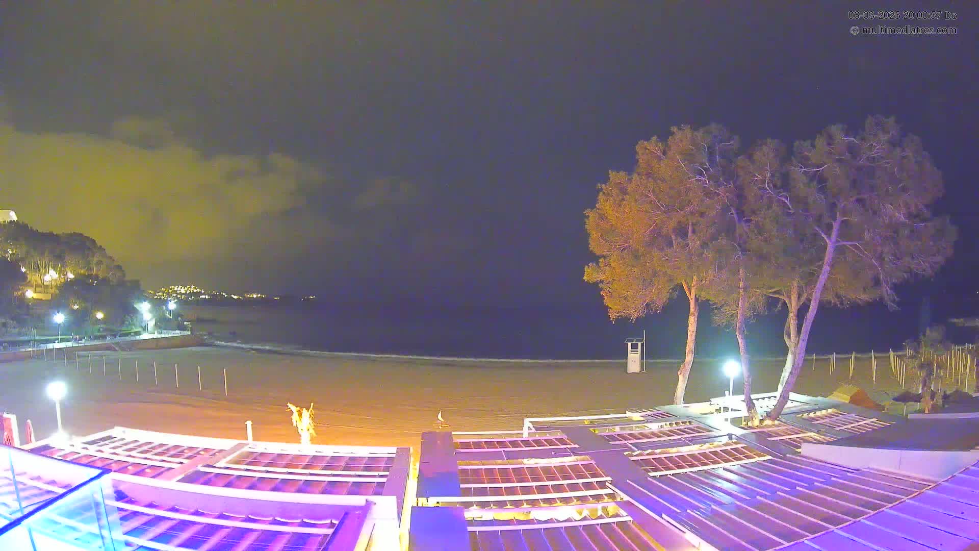 An empty beach at night under a cloudy sky is viewed from above a series of outdoor structures, with several trees standing between the structures and the shore.