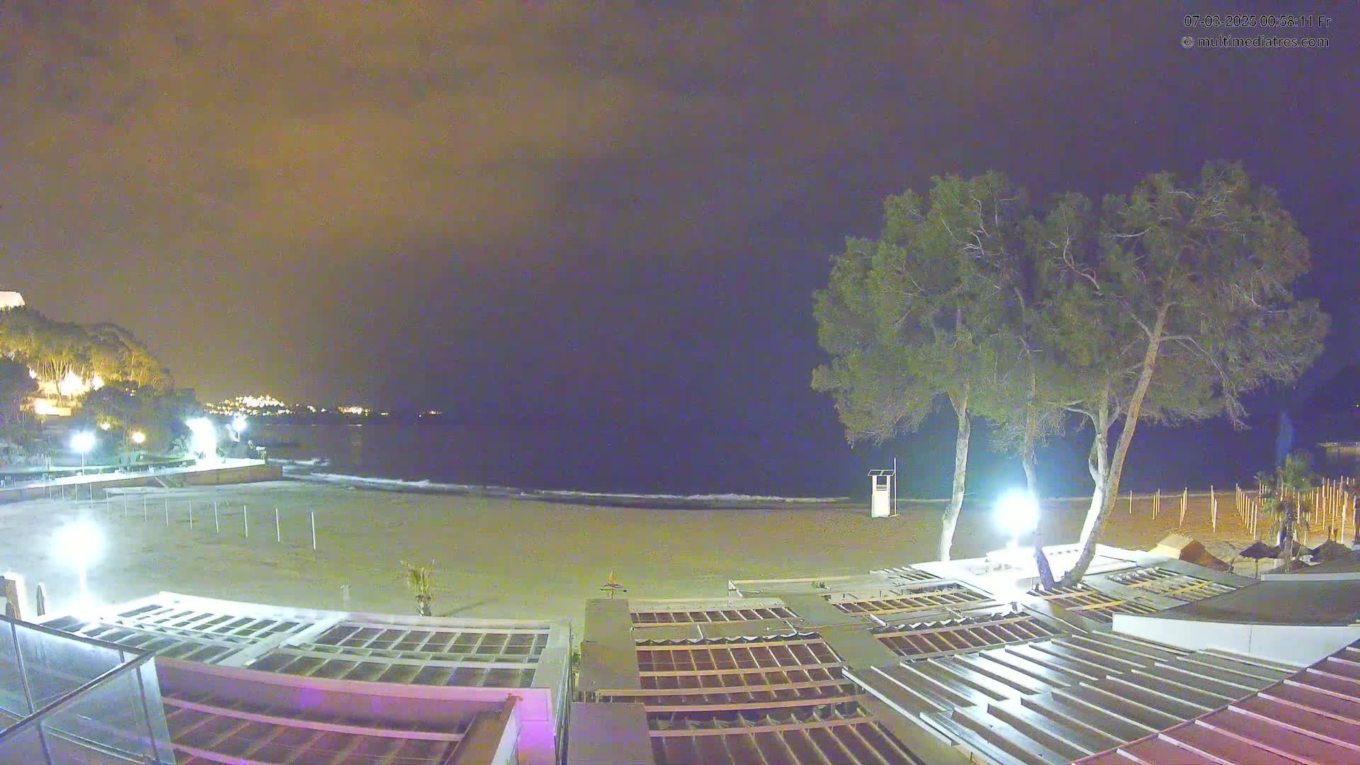 A nighttime view of a sandy beach with calm waves, illuminated by artificial lights, and two tall trees near beachside structures.