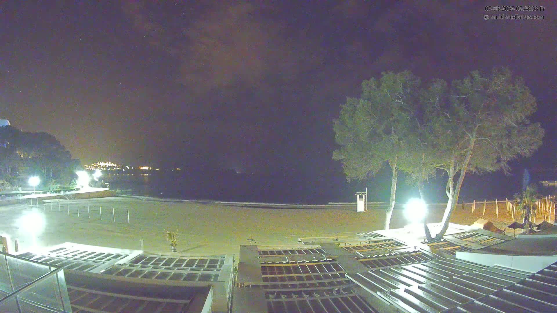 A nighttime view of a mostly empty sandy beach with structures and trees under a partly cloudy, starry sky.