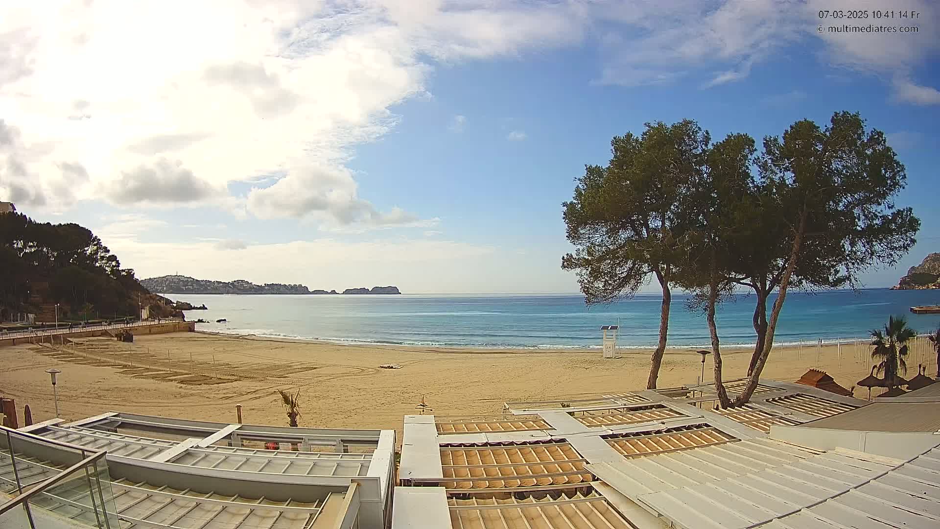 A mostly sunny day at a sandy beach with calm ocean waves, overlooking a coastline with dark green trees and distant hills.