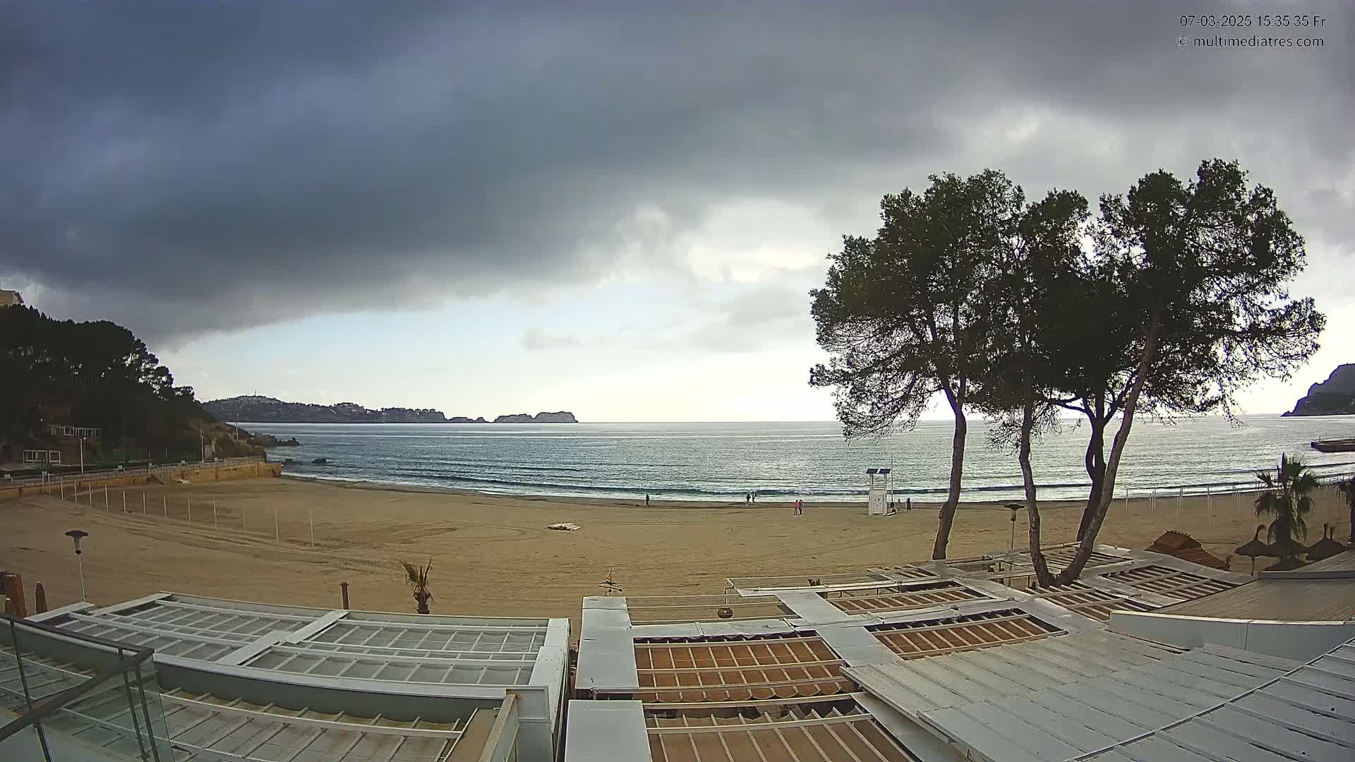 A sandy beach with calm waves under an overcast sky is viewed from above, showing beachside structures and a few people.