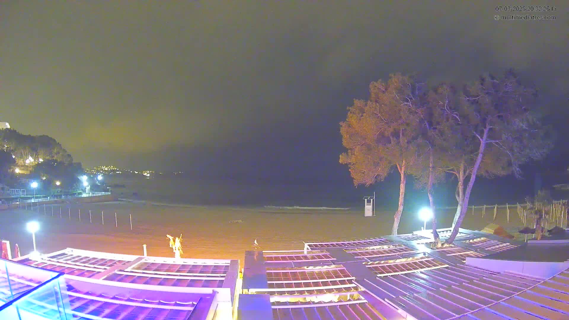 A nighttime view of a beach with calm waves, illuminated by artificial lights, featuring several trees near a beachfront structure with a series of covered areas.