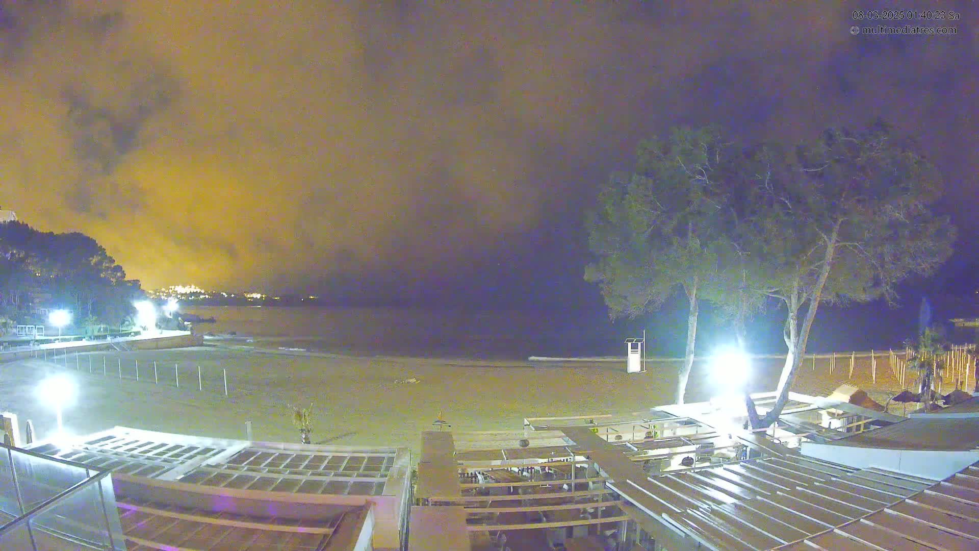 A nighttime view of a mostly empty sandy beach with a calm sea under a cloudy, yellow-tinged sky, overlooked by a beachfront restaurant and trees.
