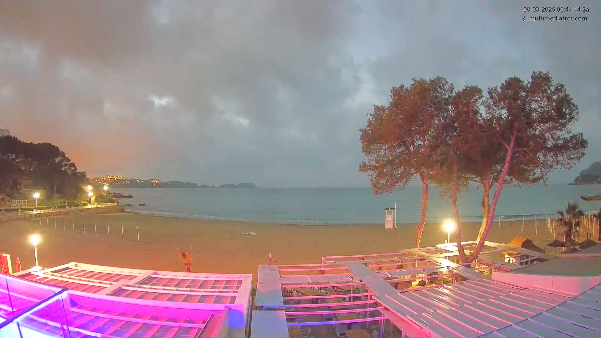 An overcast evening scene shows a sandy beach with calm water,  trees, and a partially lit beachside structure.