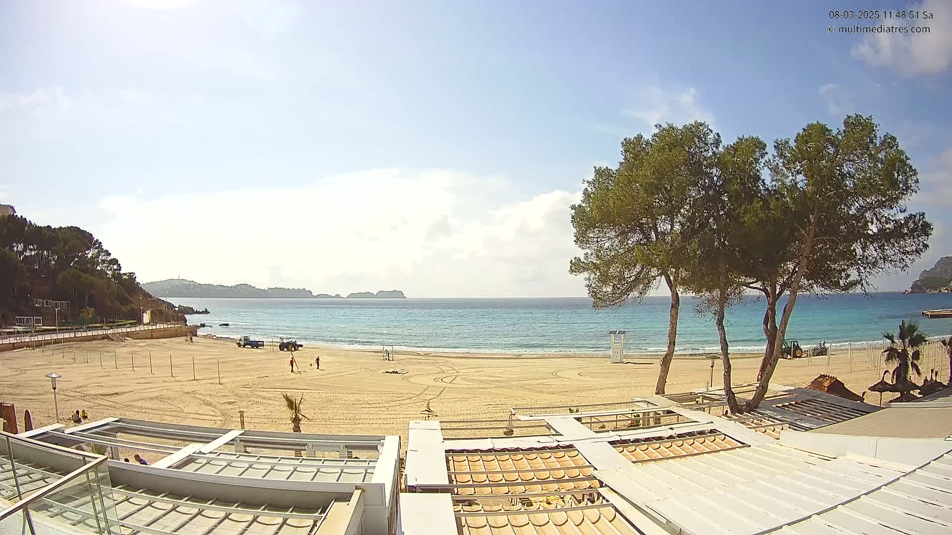 A sandy beach with calm, blue water is visible under a partly cloudy sky;  construction equipment and workers are present on the beach and some buildings are in the foreground.