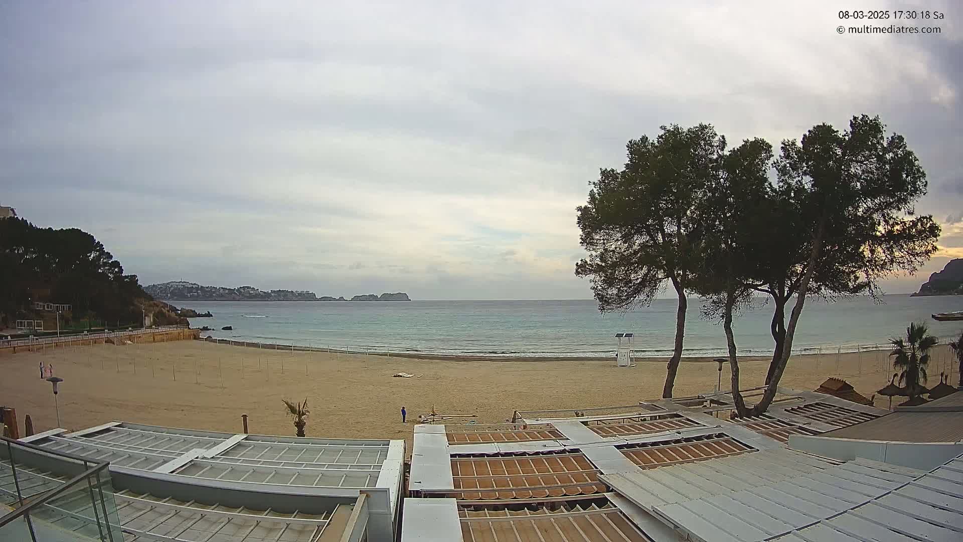 A mostly empty sandy beach with calm ocean water under an overcast sky is viewed from above, showing beach umbrellas and a lifeguard stand.