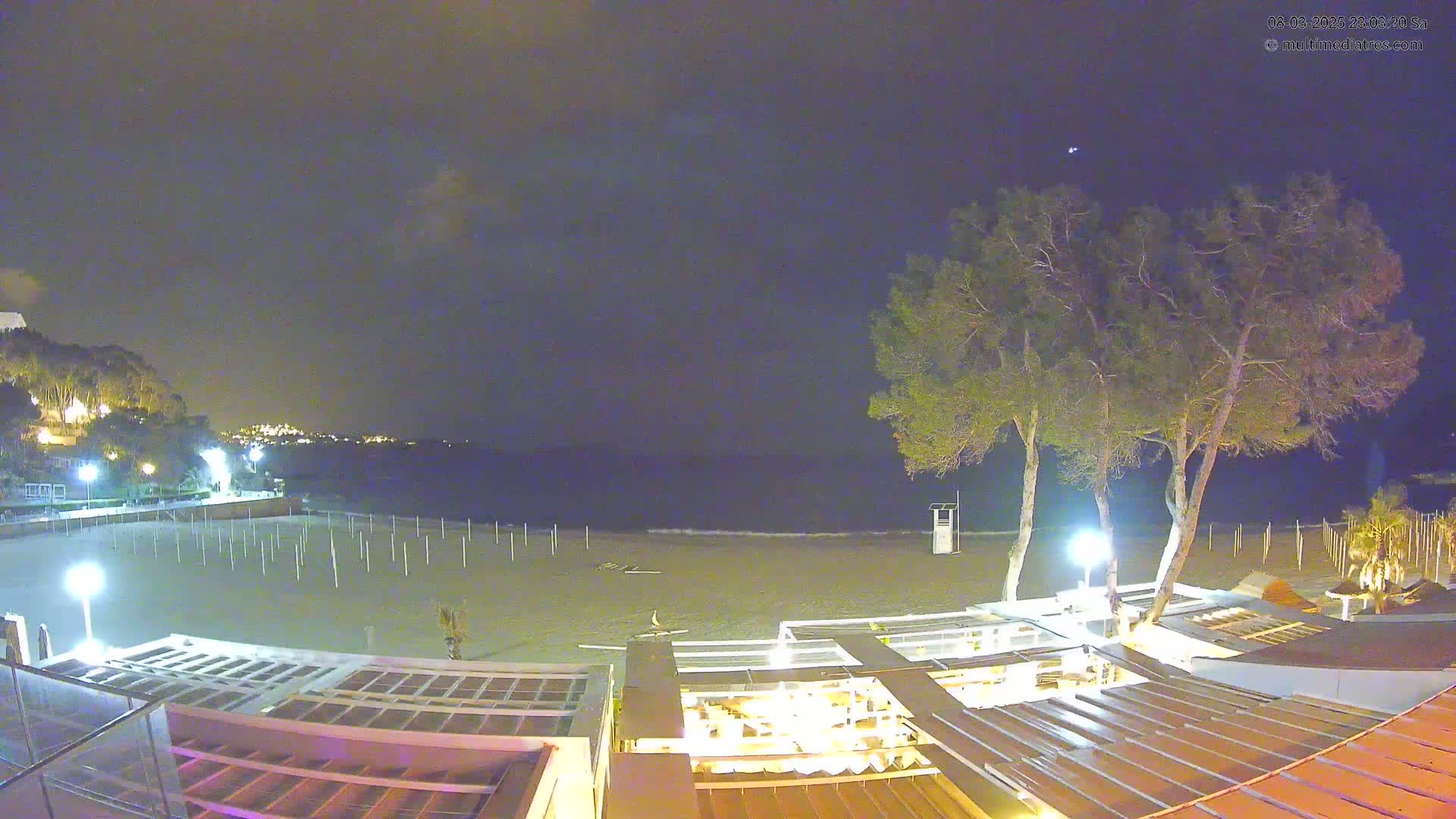 A nighttime view of a mostly empty beach with a restaurant and a few trees under a clear, dark sky.