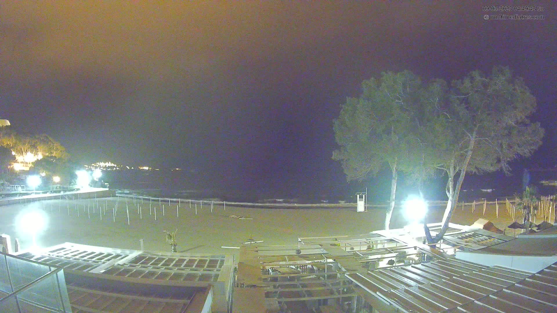 A nighttime view of an empty beach with a few lights illuminating the sand and nearby buildings, under a dark, cloudy sky.
