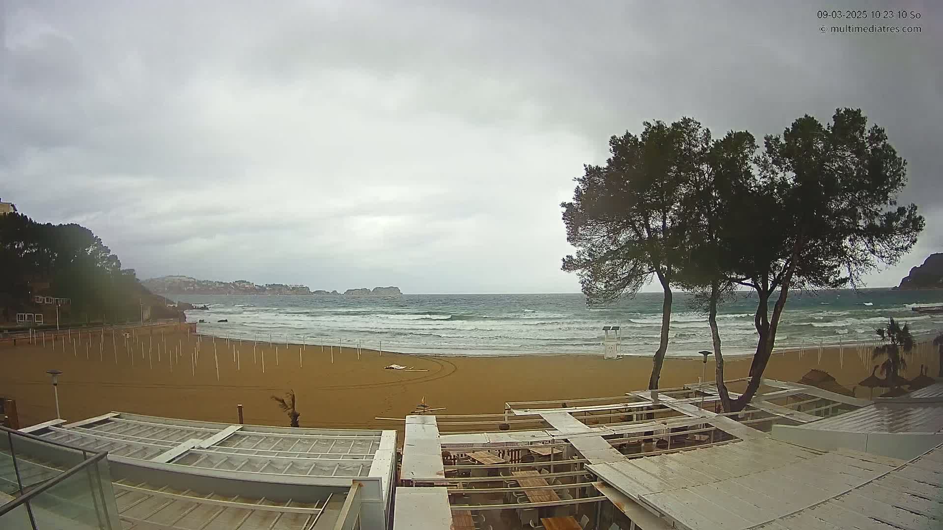 A sandy beach with choppy waves under an overcast sky, viewed from above a partially covered outdoor restaurant area near some trees.