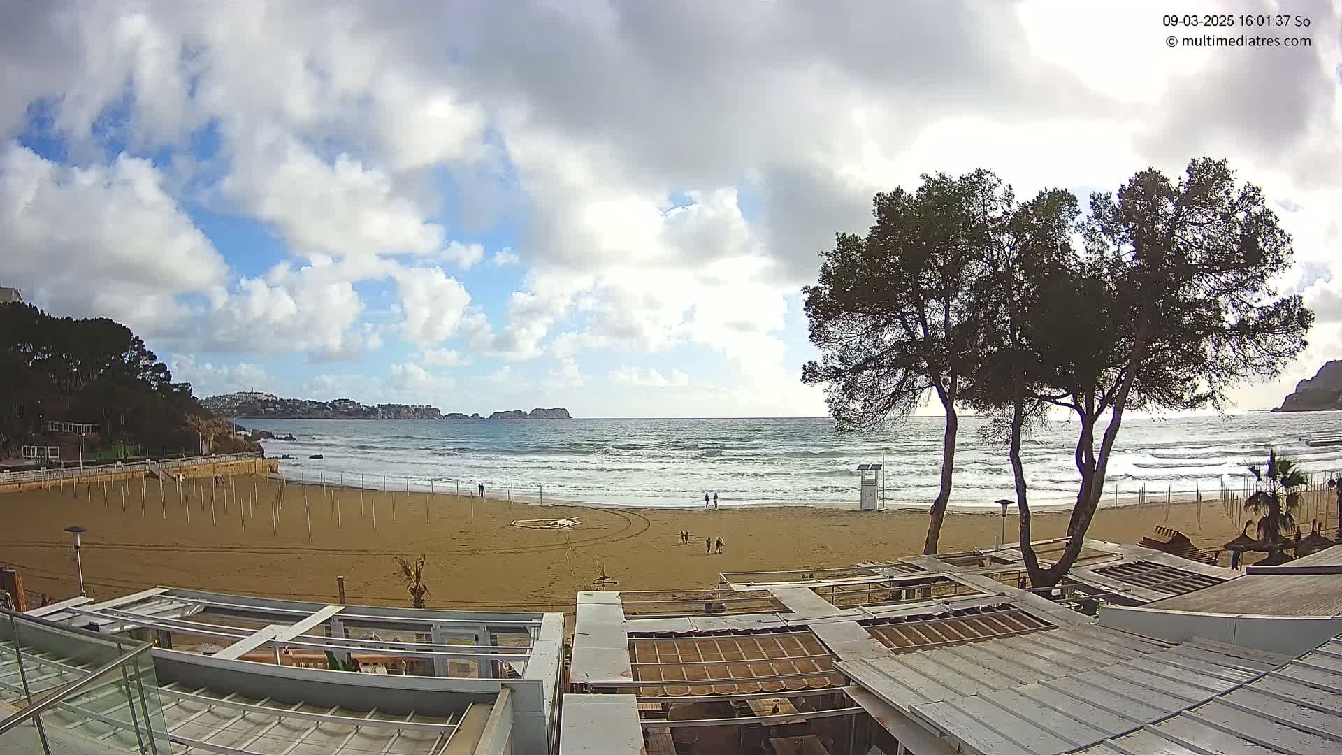 A mostly sunny day with partly cloudy skies shows a sandy beach with small waves lapping the shore, a few people walking, and several trees near beachside buildings.