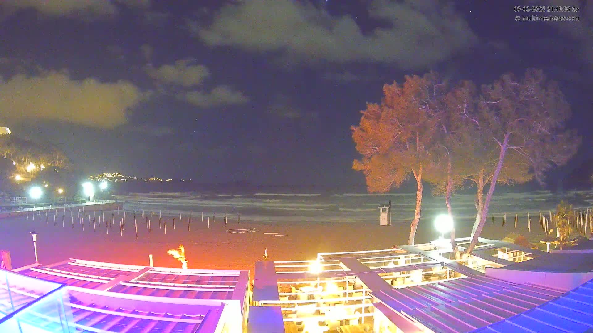 A nighttime beach scene with a partly cloudy sky shows a sandy beach with gently rolling waves, illuminated beachside structures, and several trees under the light of outdoor lamps.