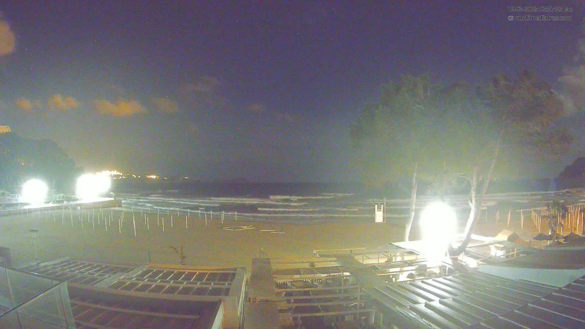 A nighttime beach scene shows a sandy shore with small waves under a partly cloudy sky, illuminated by bright lights near the beach structures.