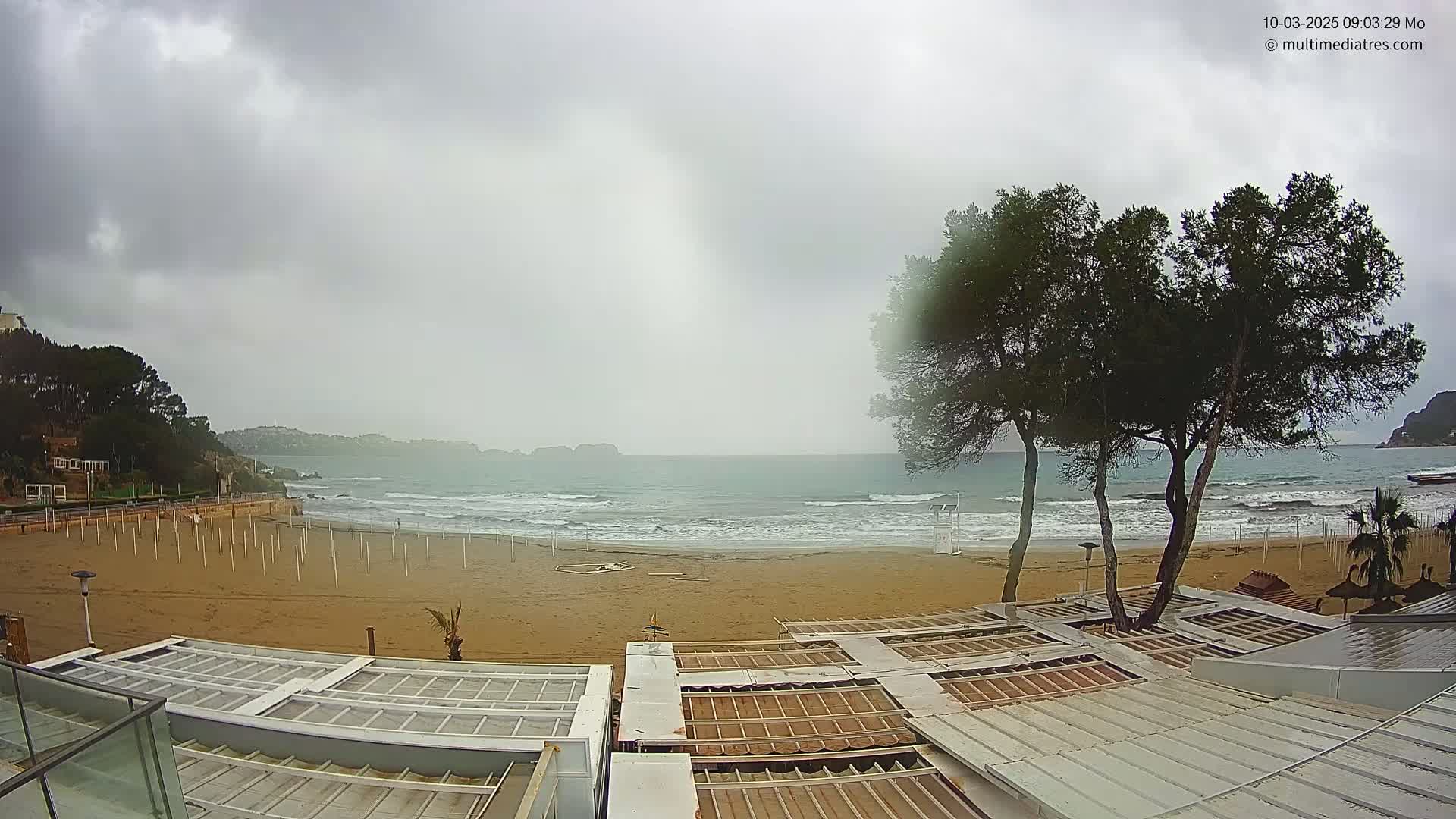 A mostly cloudy, overcast day shows a sandy beach with small waves lapping the shore, a few trees near beach umbrellas and partially covered outdoor seating areas, and hills in the distance.