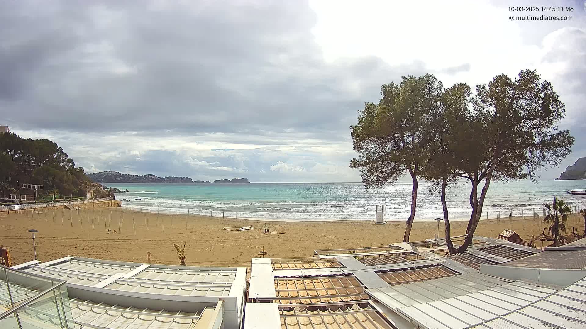 A mostly cloudy day shows a sandy beach with gentle waves lapping the shore,  backed by buildings and several pine trees.