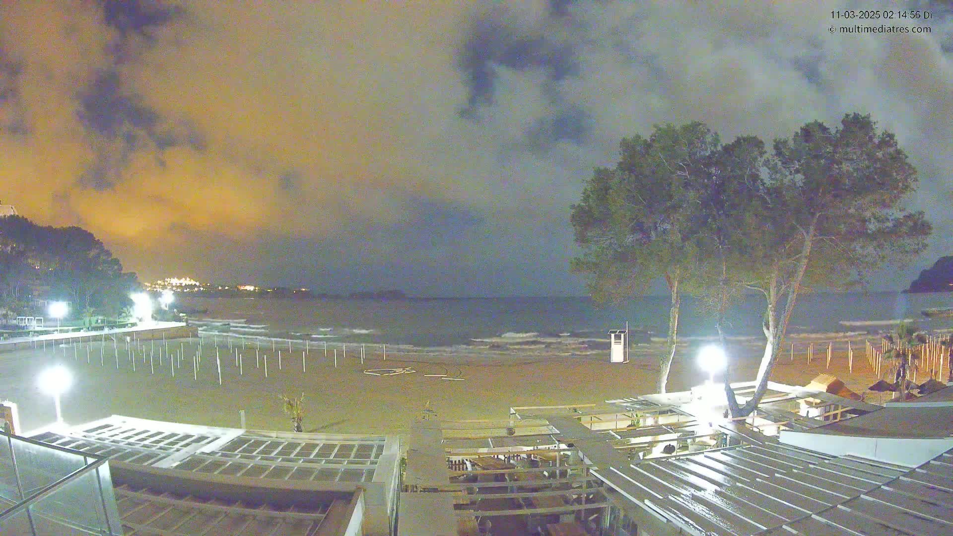 A nighttime view of a mostly empty sandy beach with calm waves, illuminated by several lights, under a cloudy sky.