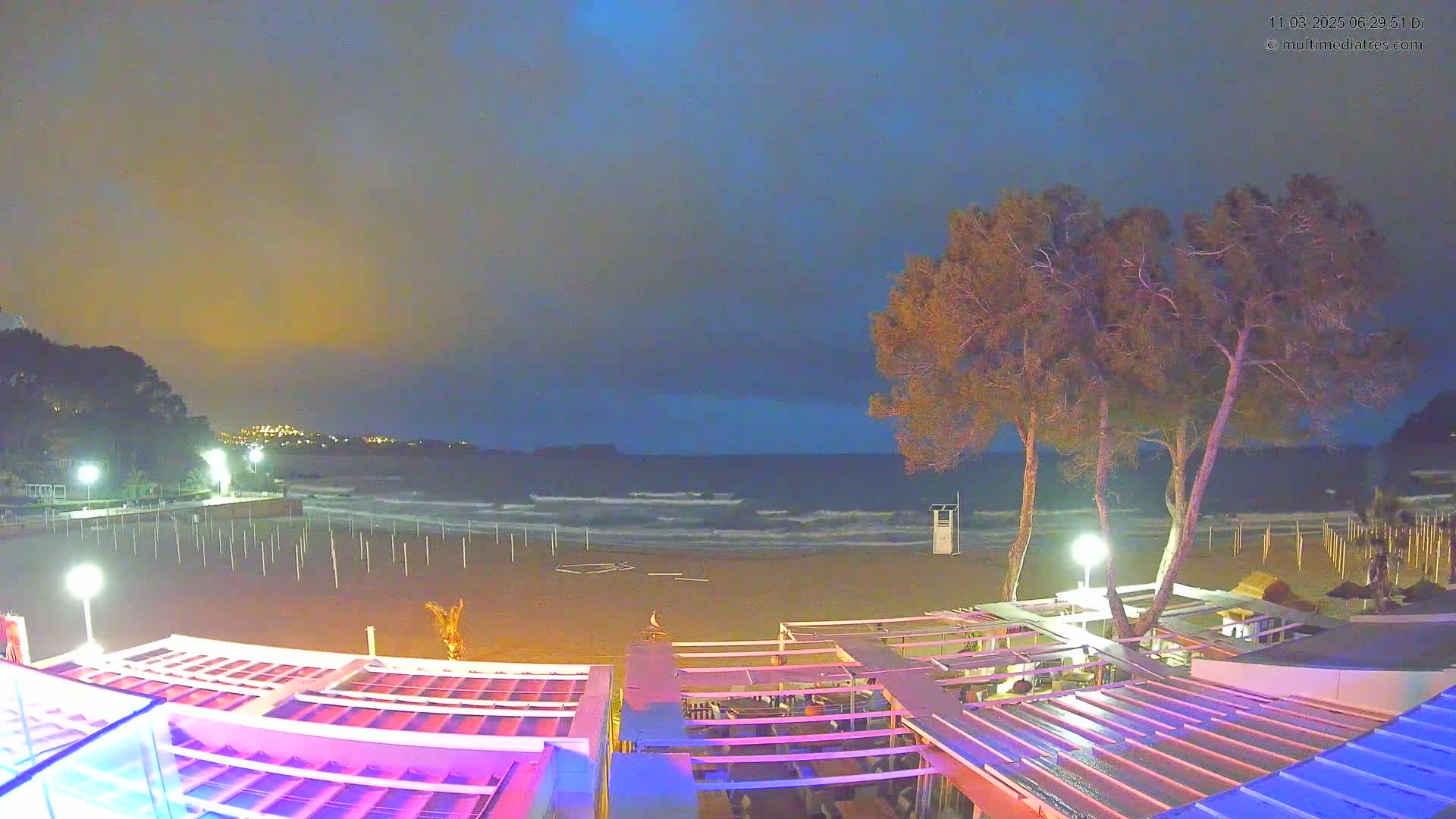 A nighttime beach scene shows a sandy shore with moderate waves under a dark blue sky, and several trees stand near beachside structures with illuminated roofs.