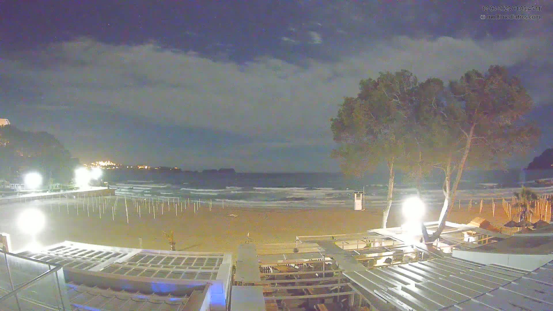 A nighttime view of a sandy beach with moderately rough waves under a partly cloudy sky, showing beachside structures and trees lit by artificial lights.
