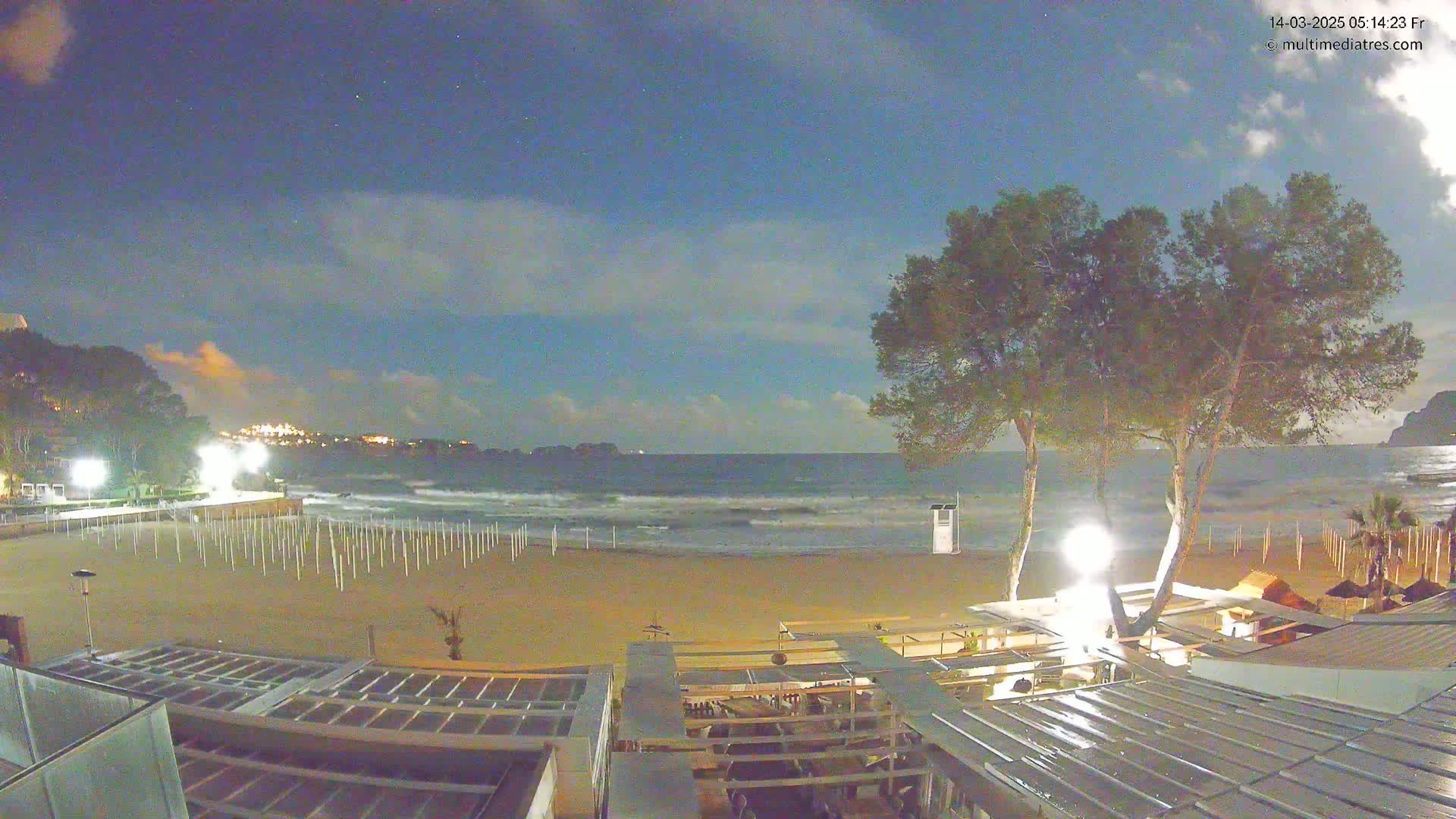 A nighttime beach scene with a mostly clear sky, gentle waves, and beachside structures illuminated under artificial light.