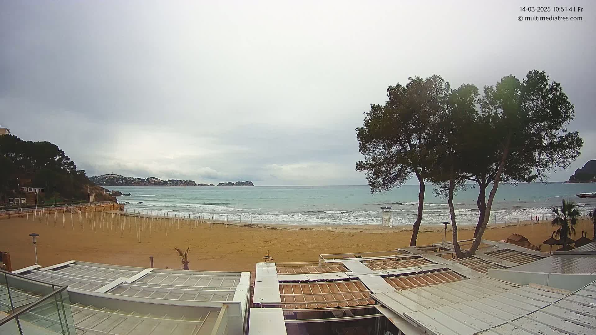 A mostly cloudy day at a sandy beach with a calm sea, featuring several trees and beach umbrellas in the foreground and buildings partially visible in the bottom right corner.