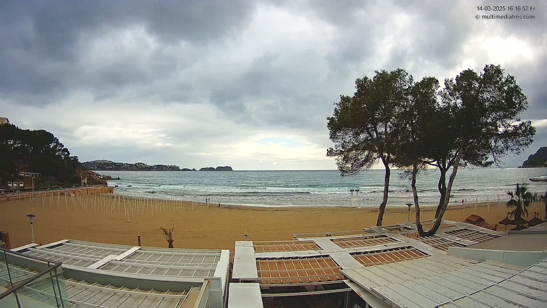 A sandy beach with calm waves under a cloudy sky is viewed from above, showing beach umbrellas and buildings in the foreground, and hills and trees in the background.