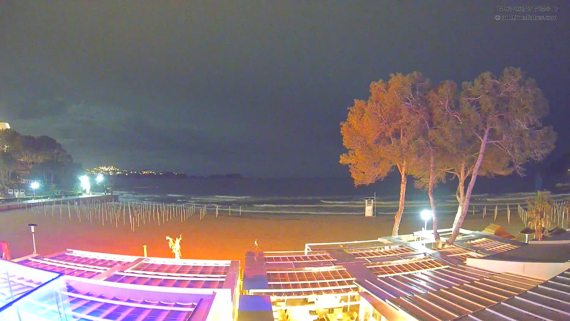 A nighttime beach scene shows a sandy shore with small waves under a dark sky, illuminated by lights from a beachfront building and distant hillside lights; several trees stand near the building.