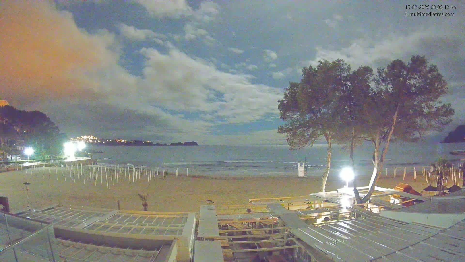 A mostly cloudy night scene shows a sandy beach with rows of beach umbrellas, a calm sea, and several trees near a beachfront building.