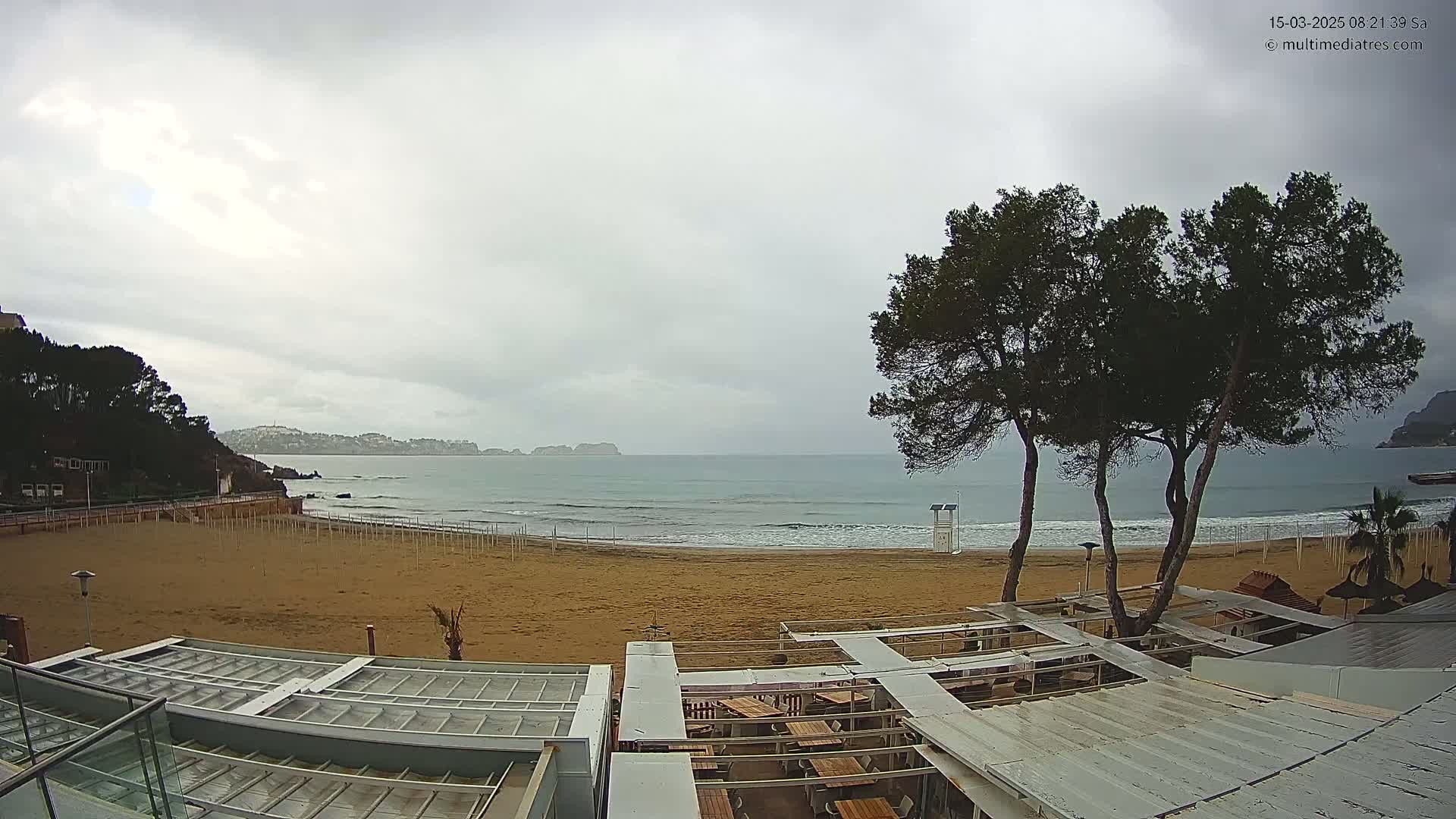A mostly cloudy day shows an empty sandy beach with calm waves, a few trees, and a mostly covered outdoor restaurant area overlooking the ocean.