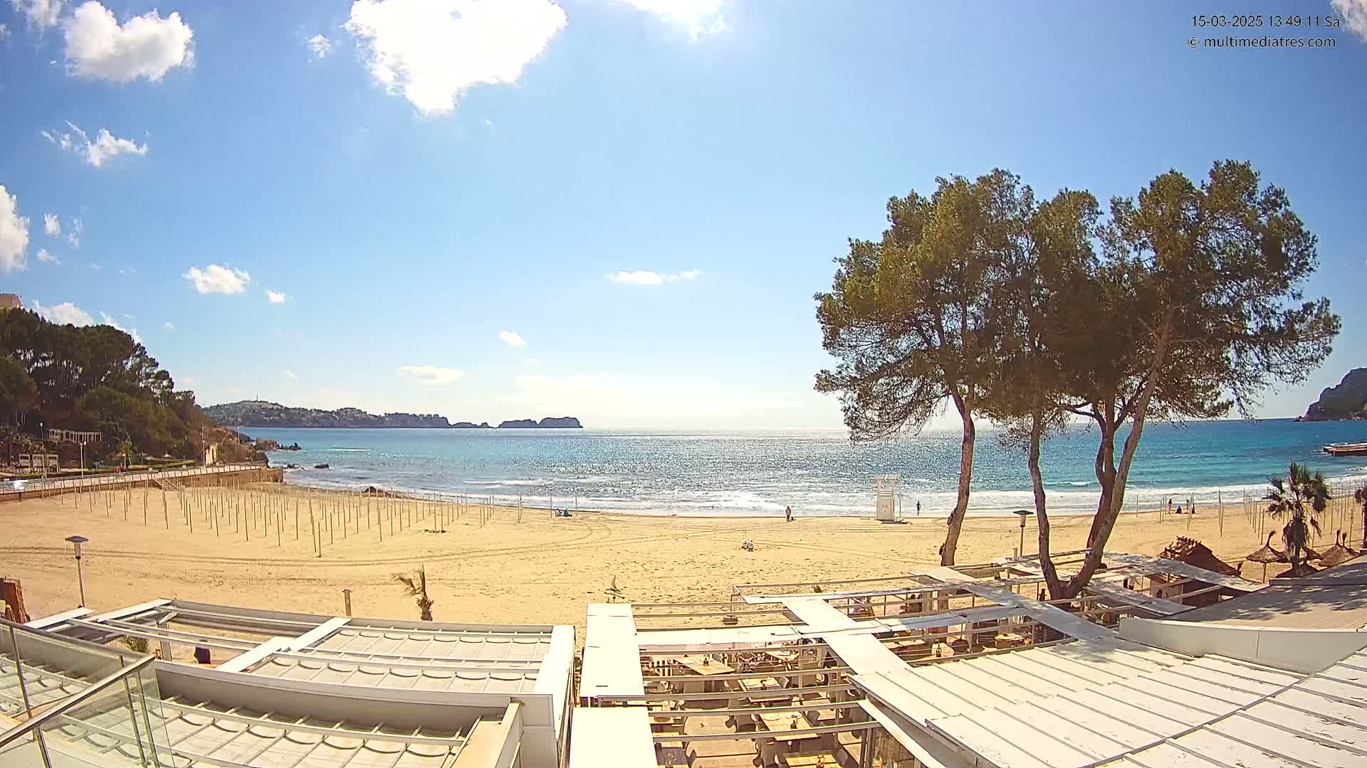 A mostly sunny day at a sandy beach with calm ocean waves, several palm trees, and a beachside restaurant with white coverings.