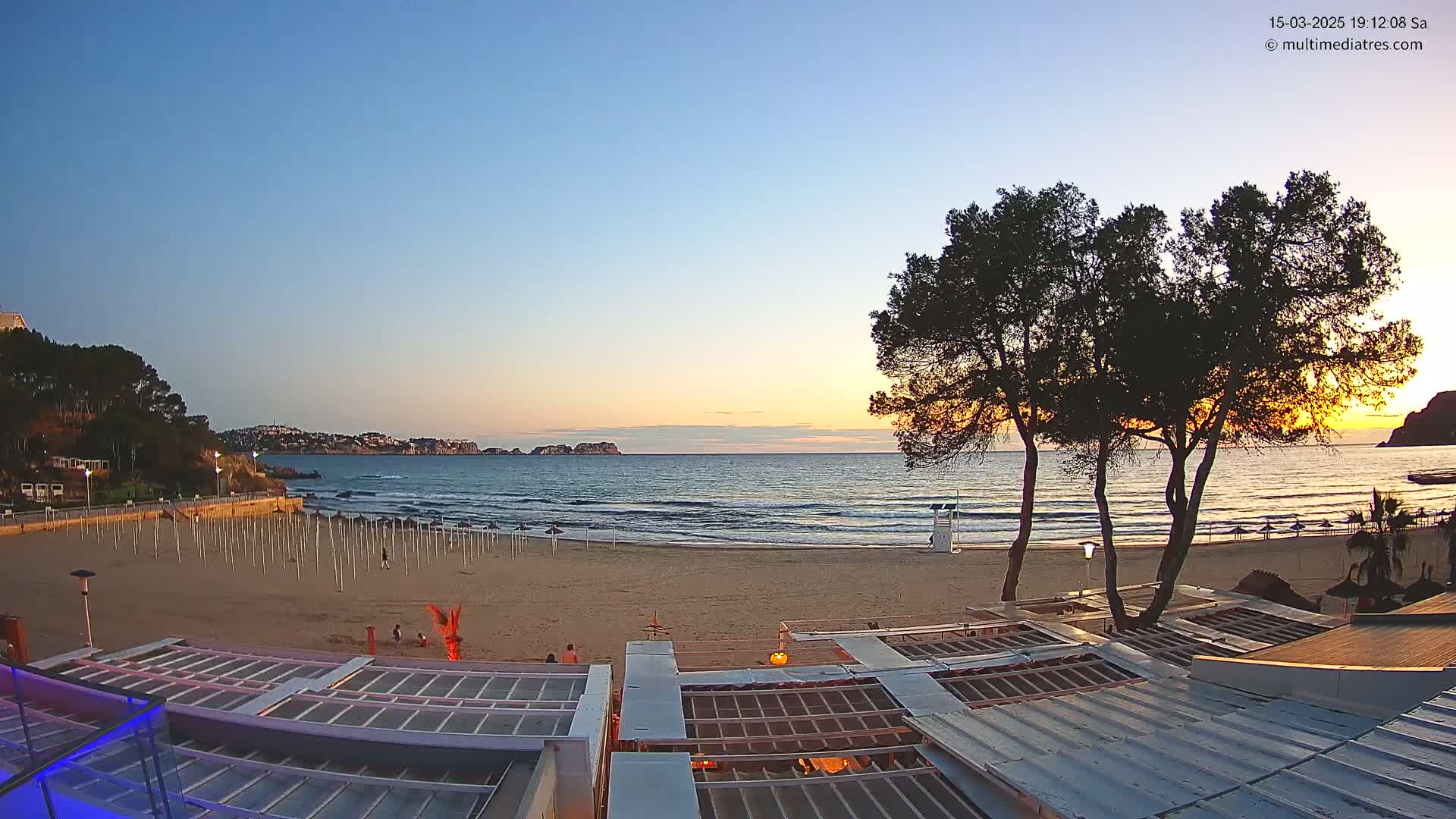 A sandy beach with calm ocean waves and a sunset over a distant coastline is viewed from atop a beachside structure.