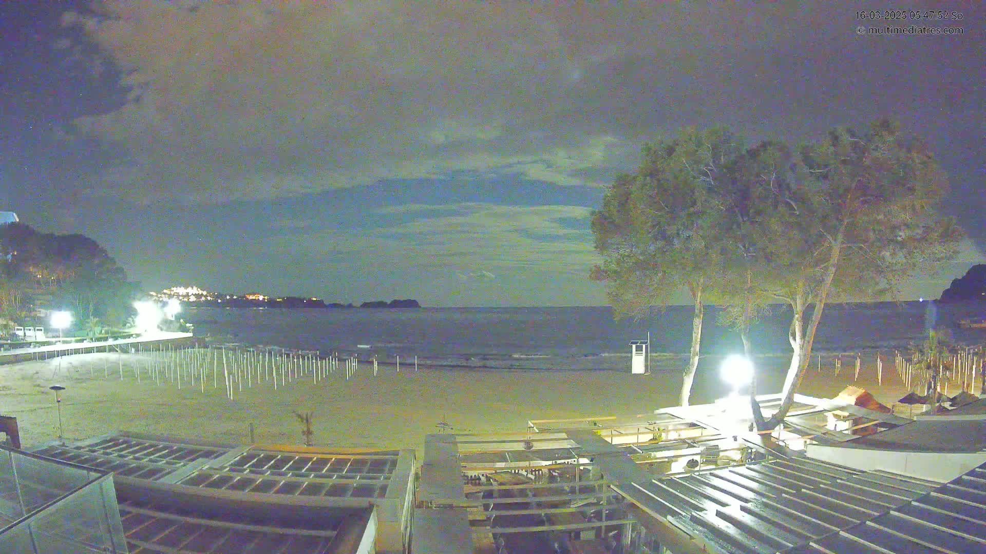 A nighttime view of a sandy beach with calm ocean waves, illuminated by artificial lights, under a cloudy sky.