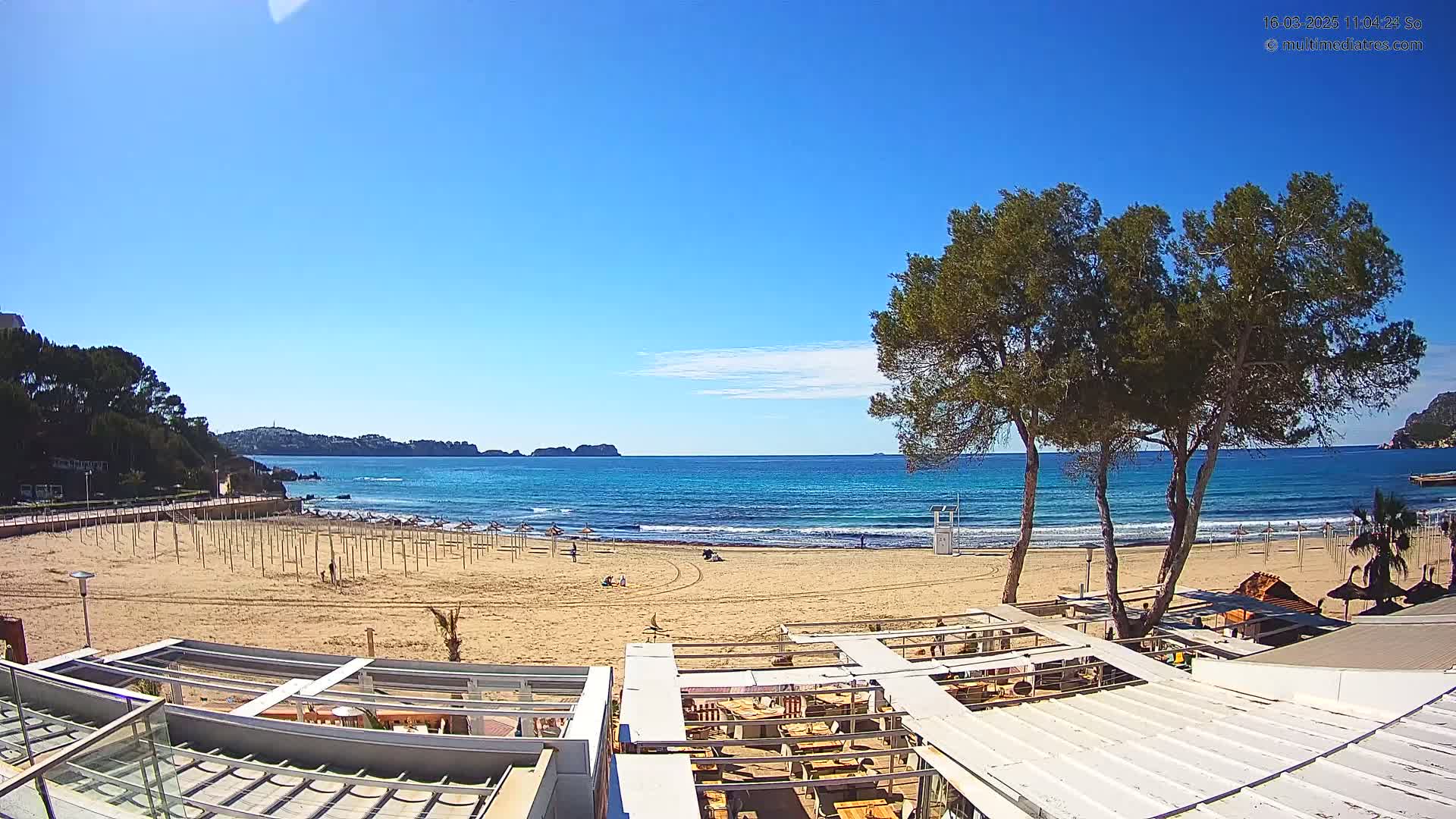 A sandy beach with calm, blue water under a clear, sunny sky is viewed from above a beachside restaurant.