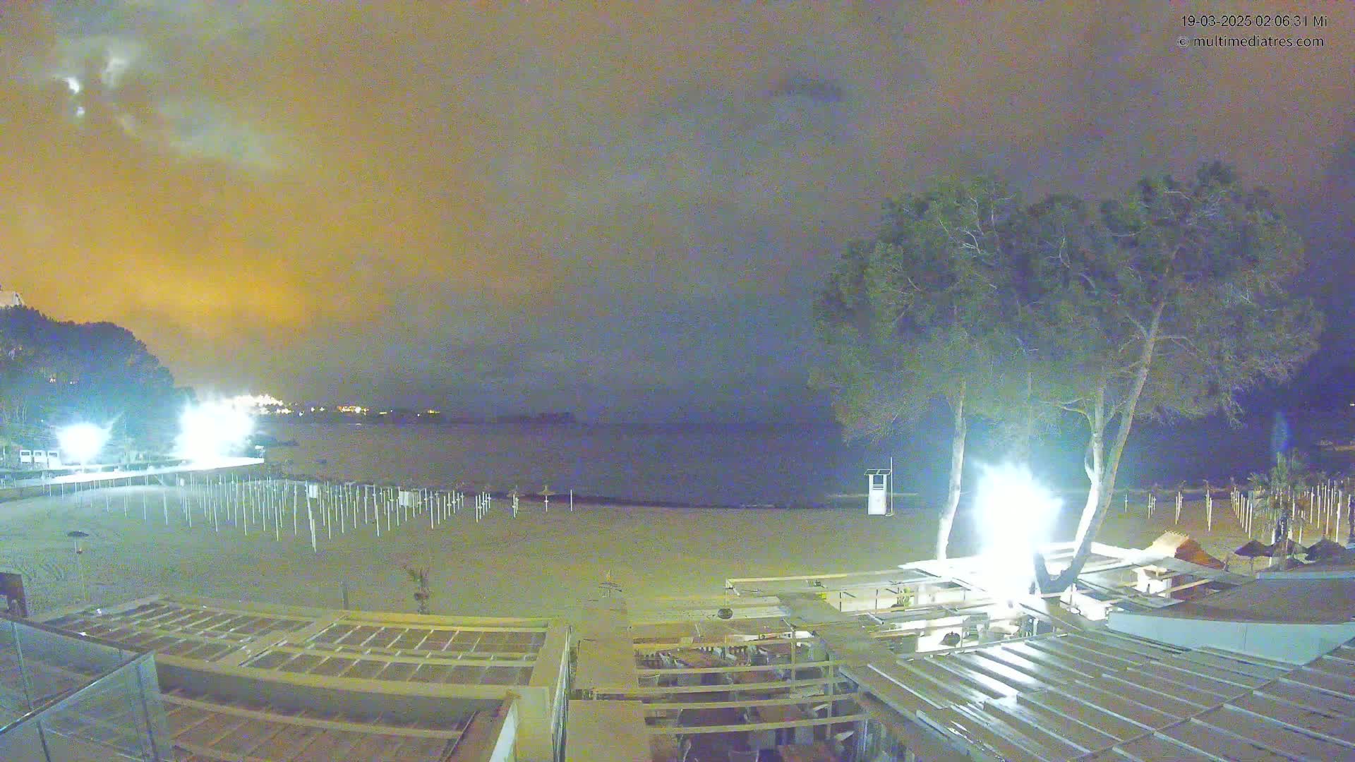 A nighttime view of a beach with a calm sea under an overcast, yellowish sky, showing beach umbrellas, trees, and a beachfront structure with bright lights.