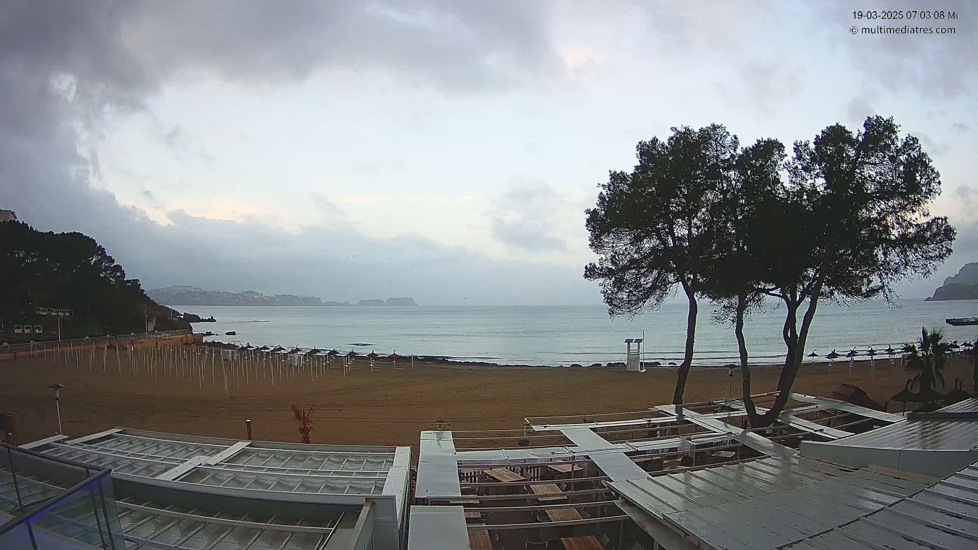 A mostly cloudy day reveals an empty sandy beach with calm ocean waves, partially obscured beach umbrellas, and a few trees overlooking a seaside restaurant.