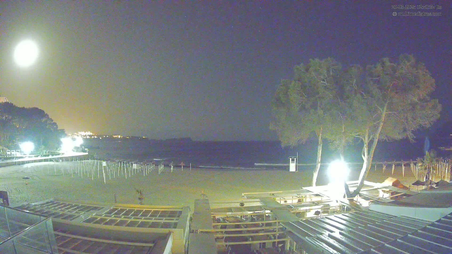 A mostly empty sandy beach at night under a bright moon is illuminated by lights near some beachside structures and trees.