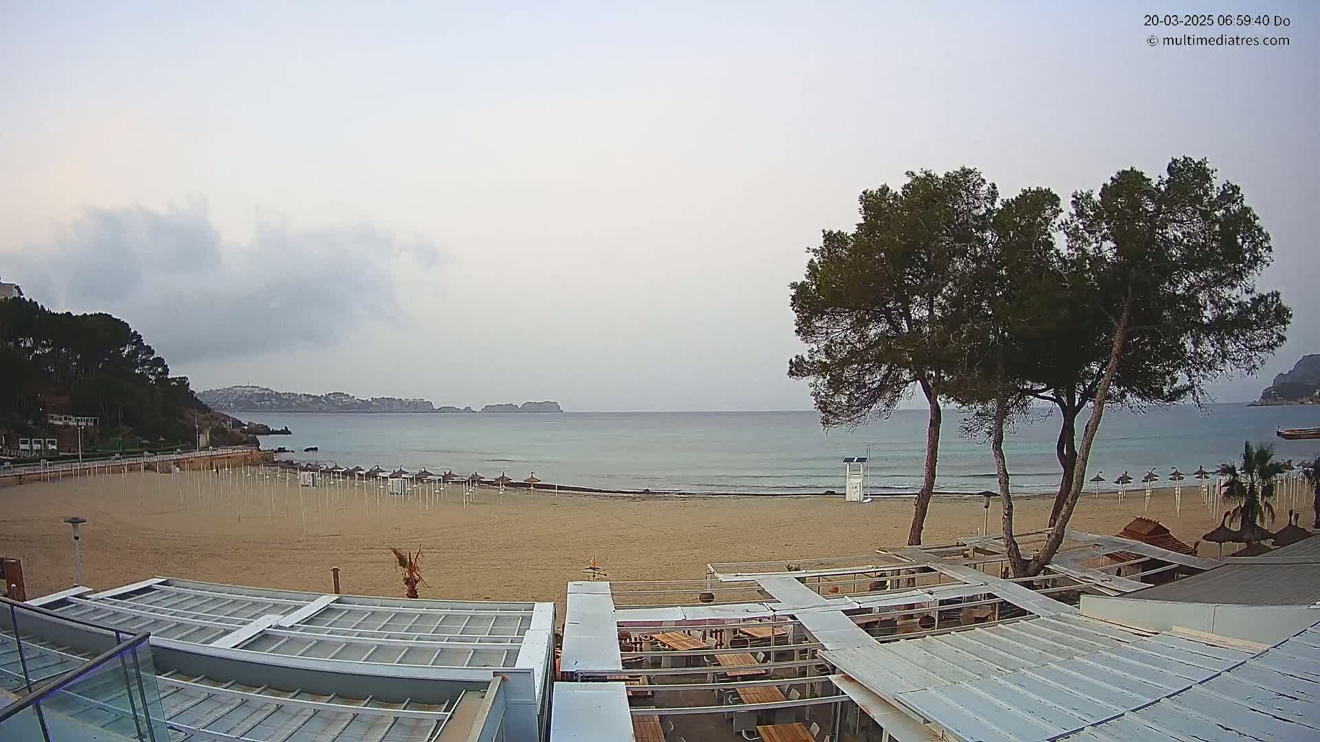 A mostly cloudy day at a sandy beach shows calm water, closed beach umbrellas, and several buildings with outdoor seating.