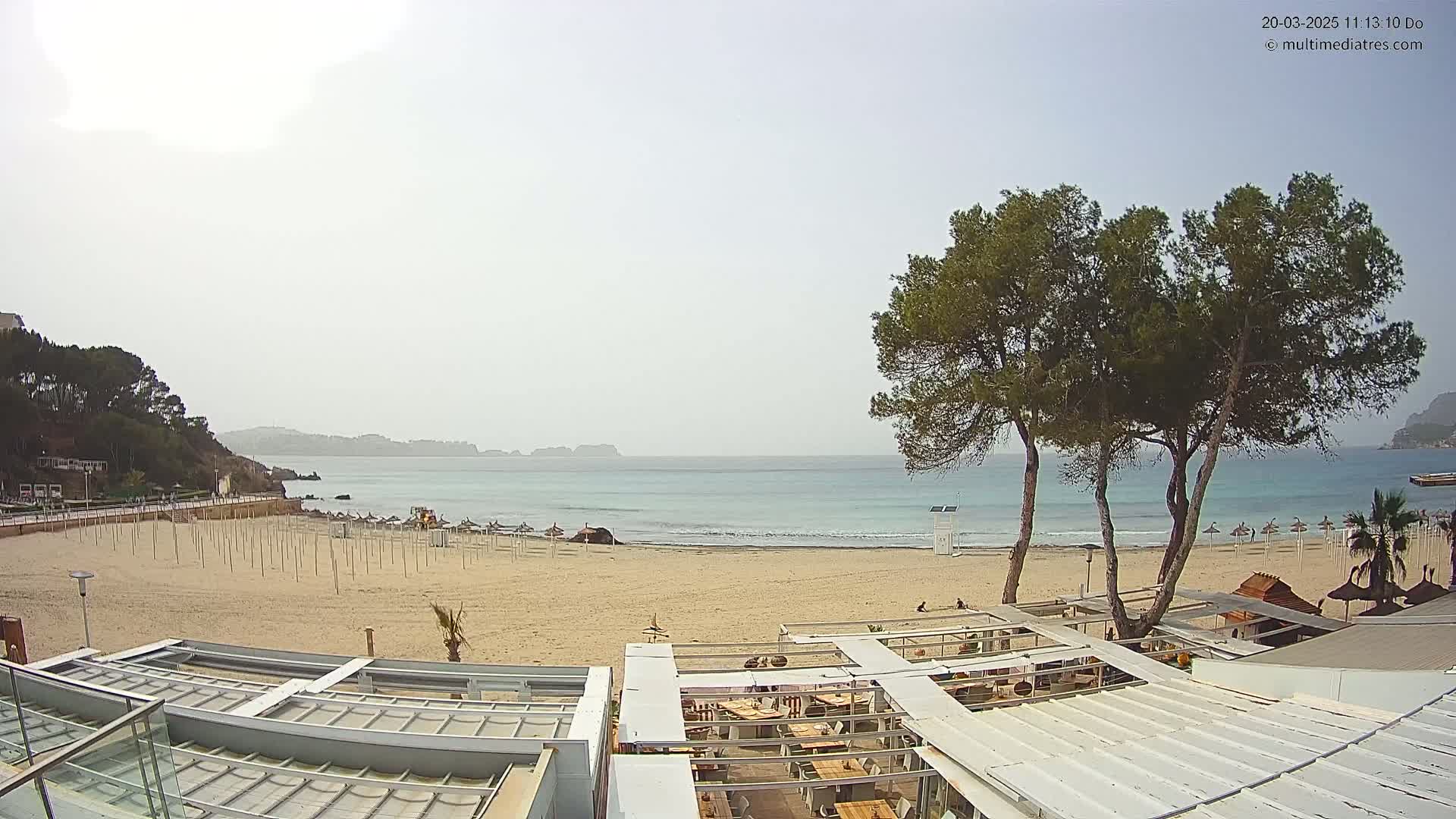 A sandy beach with calm, light-blue water, a few beach umbrellas, and several buildings with white roofs, under an overcast sky.