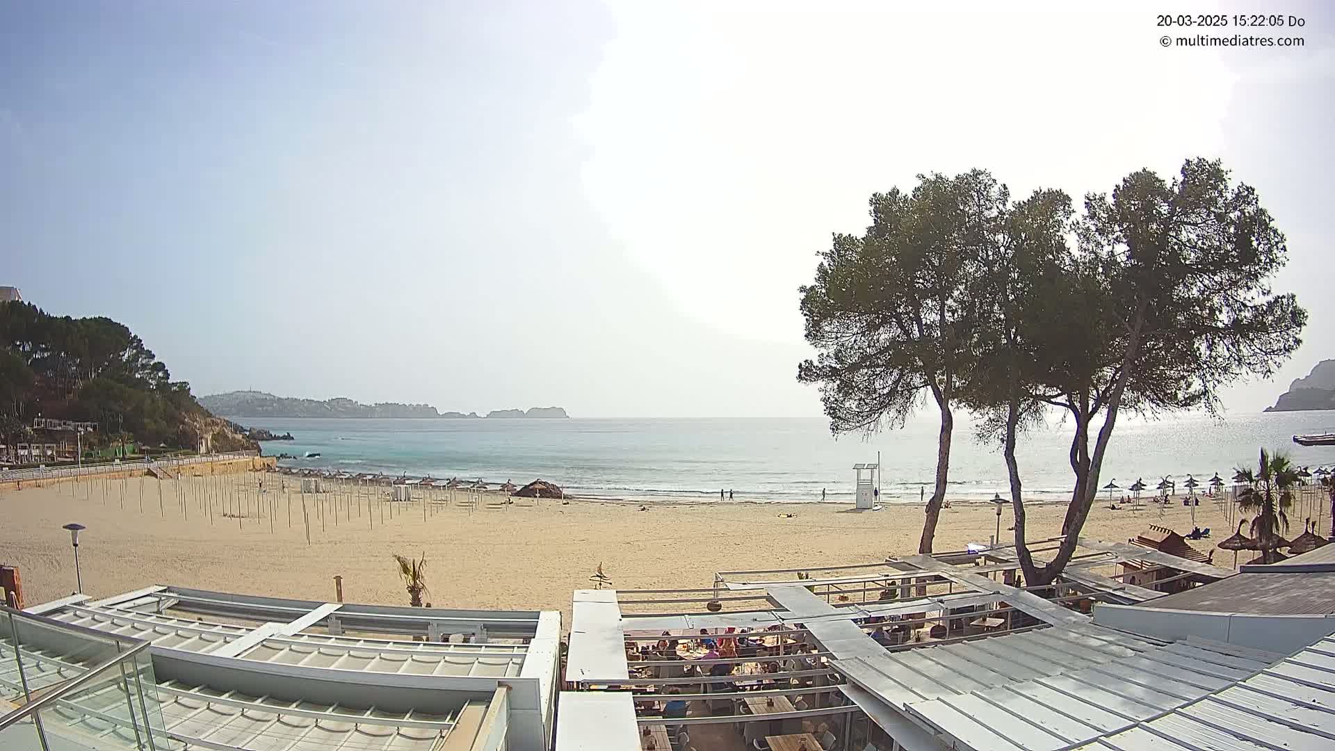 A mostly empty sandy beach with calm, light blue water stretches to a distant coastline under a clear, bright sky, and overlooks a partially covered outdoor restaurant.