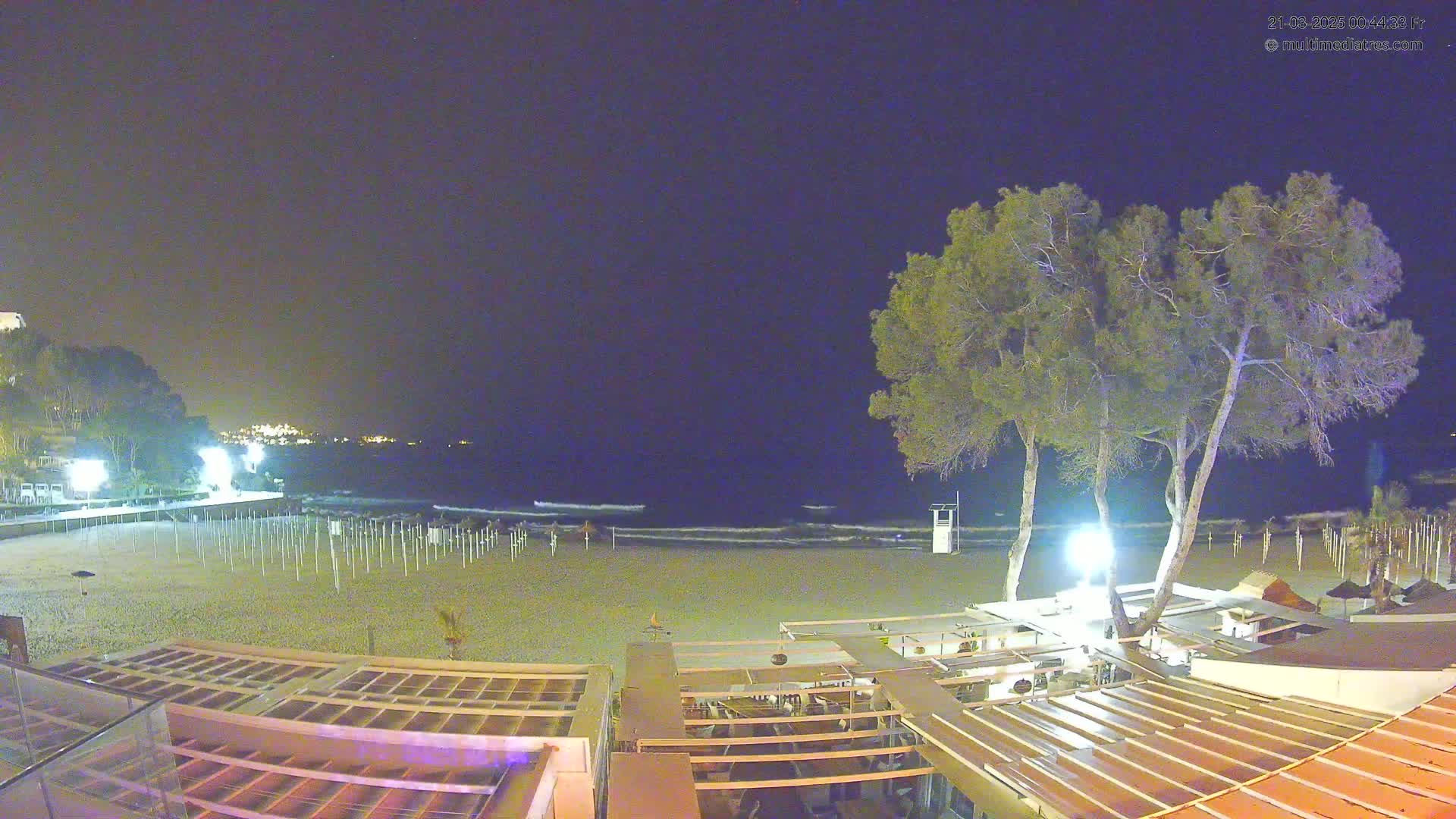 A nighttime view of a beach with calm waves, illuminated beach umbrellas, several trees, and a beachfront restaurant under a clear, dark sky.
