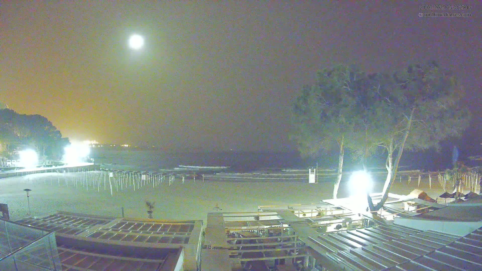 A nighttime beach scene under a full moon shows a mostly empty sandy beach with rows of closed beach umbrellas, a few buildings, and some trees illuminated by lights.