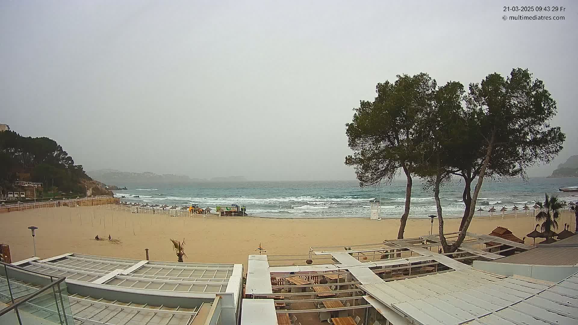 A sandy beach with some umbrellas and a few people is visible under an overcast sky, with moderate waves crashing on the shore near a beachside restaurant.