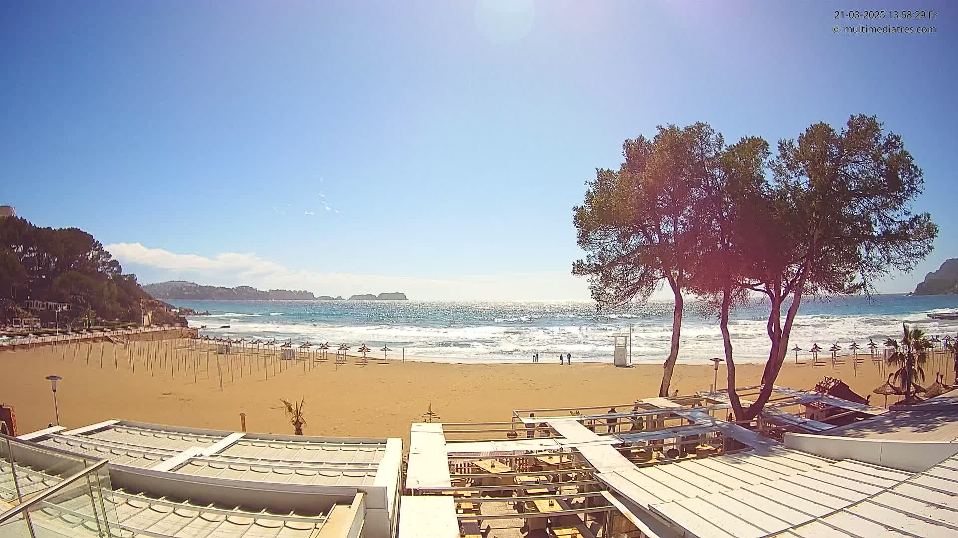 A sandy beach with calm waves, closed umbrellas, and a few people visible in the distance under a sunny sky, overlooked by a partially covered outdoor restaurant area and several pine trees.