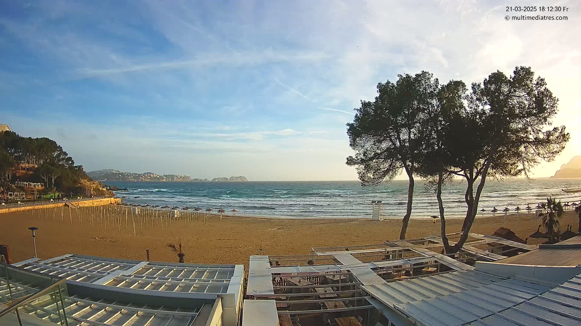 A sandy beach with moderate waves under a mostly clear sky, viewed from a beachfront establishment with several trees in the foreground.