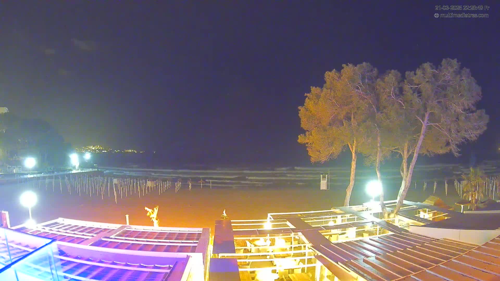 A nighttime view of a sandy beach with gentle waves under a mostly clear sky, showing an outdoor restaurant area with lights and several trees nearby.