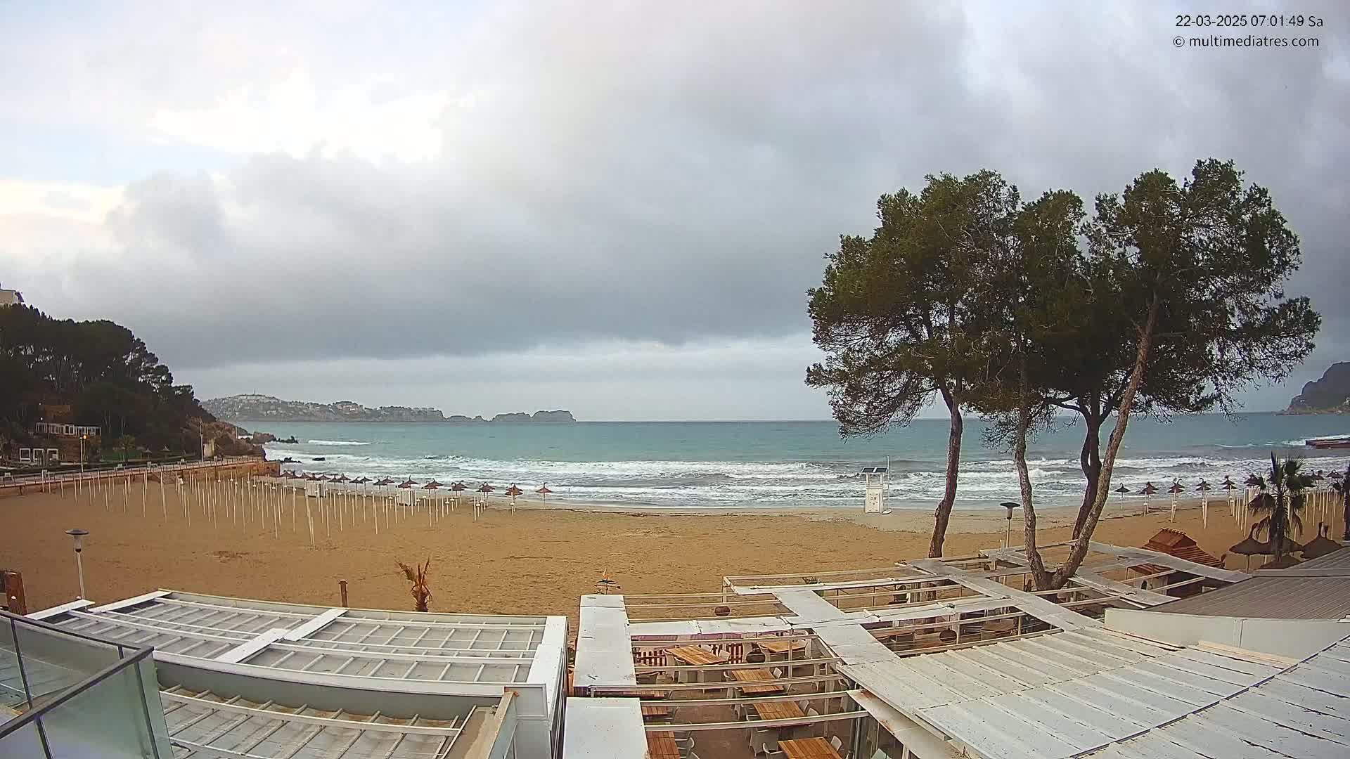 A mostly cloudy day at a sandy beach with some waves, featuring several closed beach umbrellas and a beachside restaurant with outdoor seating.